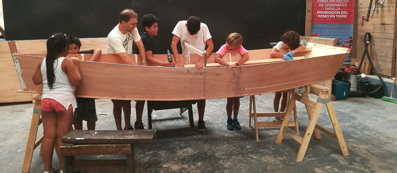 Chicos y docentes alrededor del casco de un bote de madera en construcción, apoyado sobre caballetes en el galpón.