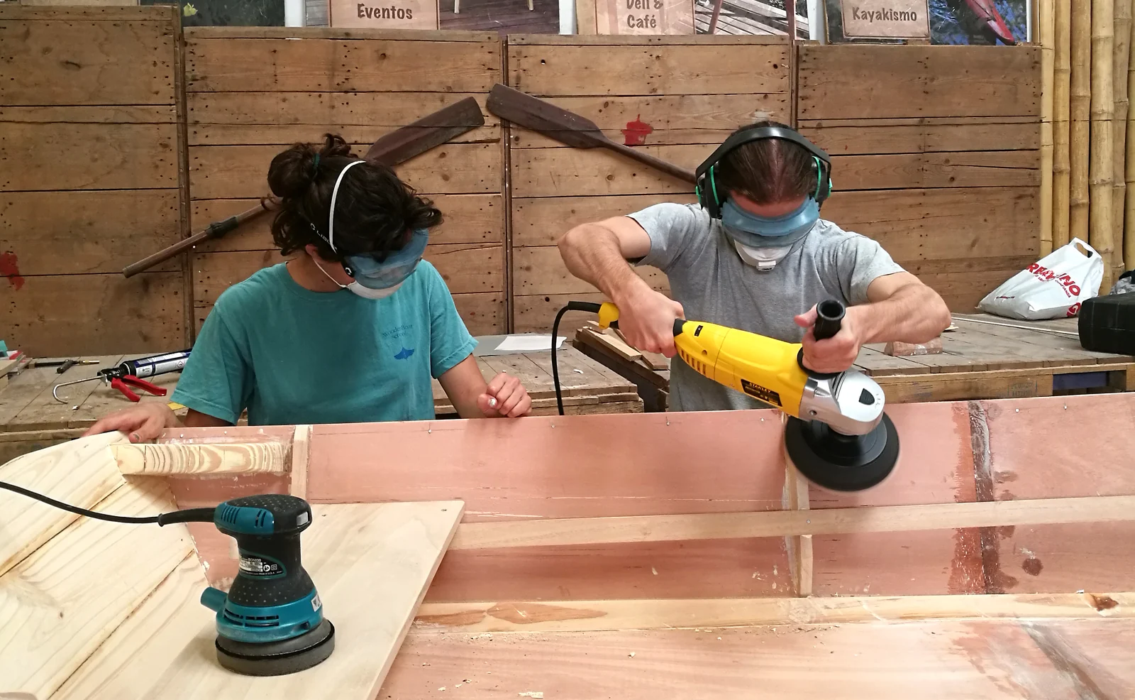 Dos chicos con protectores auditivos y barbijo lijan una tabla junto al casco del bote.